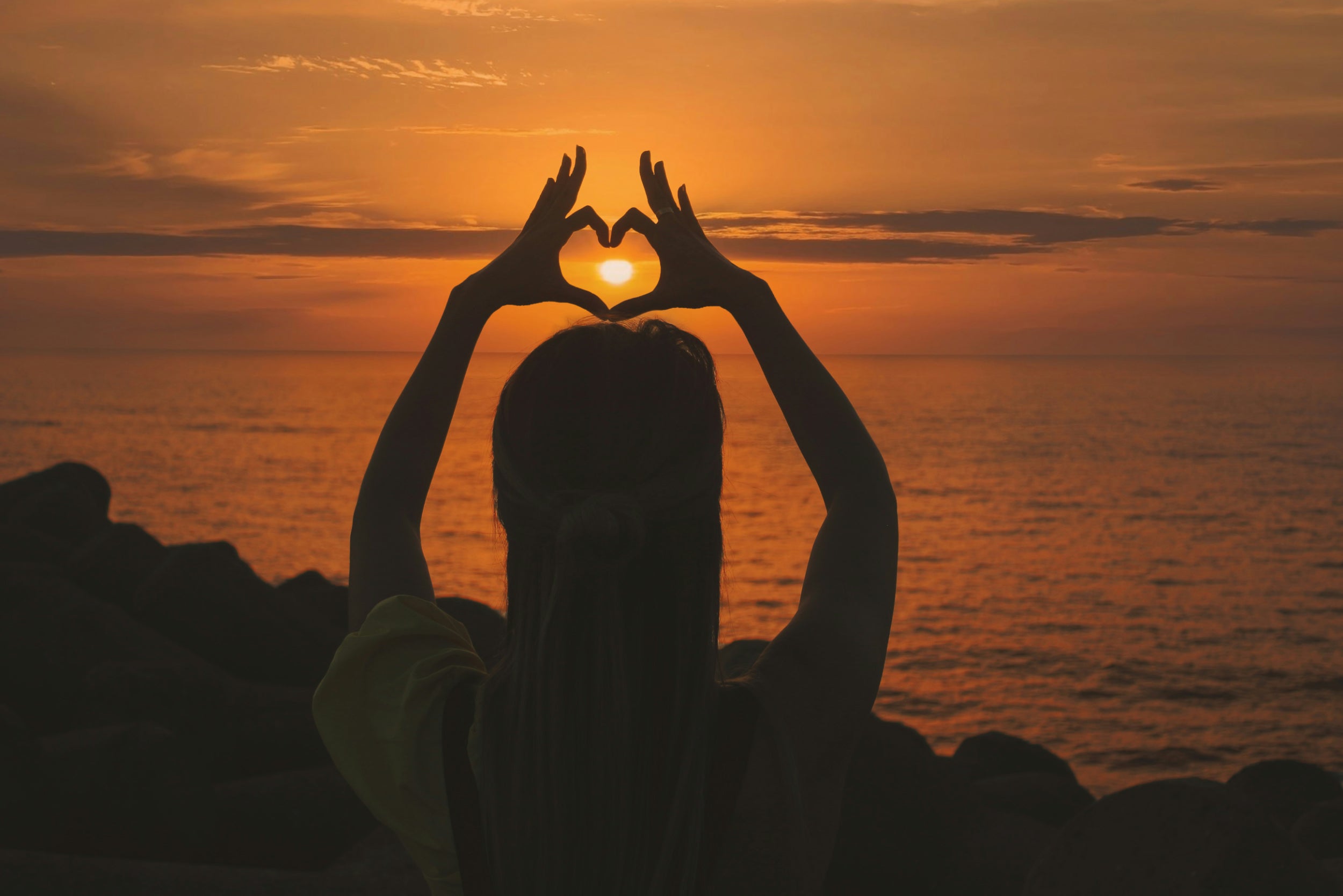 Photograph of woman holding up hands in a heart shape in front of a sunset